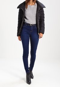 Zwarte pufferjas met een hoge kraag, grijze geribbelde trui, donkere blauwe skinny jeans en zwarte enkellaarzen. Casual outfit.