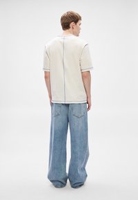 Camisa de cor creme com detalhe em costura azul nas costas, combinada com jeans de ganga azul-claro de perna larga. Textura do tecido suave visível.