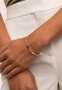 Bracciale in oro con perline multicolori in turchese, rosa e trasparente, con un piccolo ciondolo circolare, indossato su un polso.