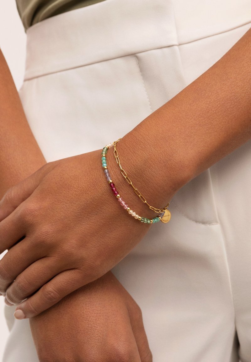 Bracciale in oro con perline multicolori in turchese, rosa e trasparente, con un piccolo ciondolo circolare, indossato su un polso.