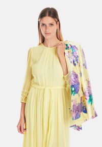 Gelbes plissiertes Kleid mit tailliertem Schnitt; kombiniert mit einer floral gemusterten Jacke mit lila und grünen Akzenten. Die Bluse hat durchsichtige Ärmel.