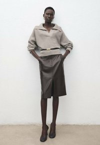 Maglione grigio chiaro lavorato a maglia con un ampio colletto, abbinato a una gonna in pelle marrone scuro con spacco frontale, e tacchi neri.