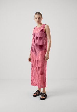 Rosa, ärmlös klänning med ett meshdesign som har cirkulära utskärningar. Modellen bär svarta sandaler med remmar. Slät textur, knälång.
