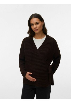 Ung kvinne med mørkt hår iført en mørkebrun innhyllet genser over en hvit skjorte, som forsiktig holder magen mens hun står mot en ensfarget bakgrunn.