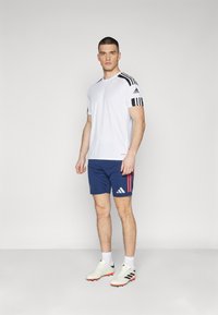 Biele atletické tričko s čiernymi pruhmi na pleciach, skombinované s tmavomodrými šortkami s červenými akcentmi a bielymi teniskami.