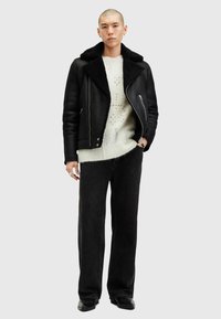 Schwarze, mit Schaffell gefütterte Lederjacke, cremefarbener Strickpullover mit Mustern, schwarze weit geschnittene Jeans und schwarze Schuhe. Minimalistisches Design mit klaren Linien.