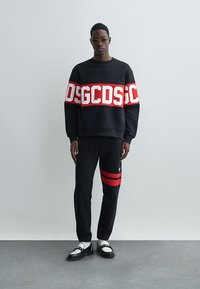 Čierna mikina s bielym a červeným nápisom, v kombinácii s čiernymi teplákmi so zlatými pruhmi. Topánky sú bielo-čierne s dekoračnými prvkami.