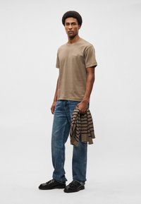 Khaki színű, rövid ujjú póló kis mellzsebbel, kék, egyenes szabású farmernadrággal párosítva, valamint egy barna csíkos kötött pulóverrel. Fekete cipő.