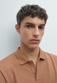 Young man with short curly dark hair wearing a brown collared shirt, looking directly ahead against a plain light background.