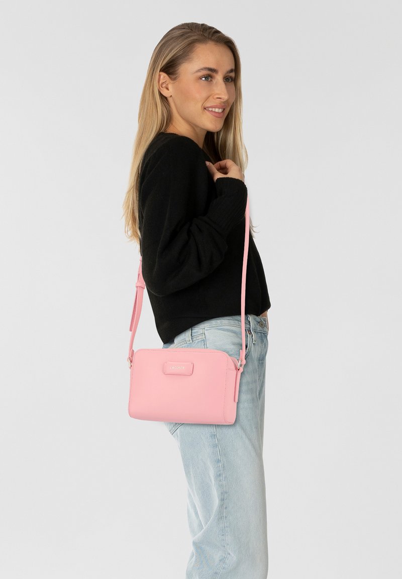 Borsa a tracolla rosa con una texture in pelle liscia, forma rettangolare, dotata di un logo frontale e di una tracolla regolabile.