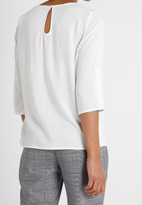 Person som bär en vit blus med detalj i form av ett nyckelhål i ryggen och grå rutiga byxor, stående mot en enfärgad ljus bakgrund.