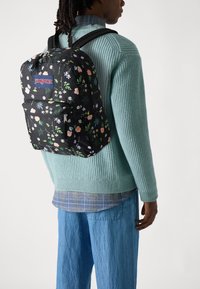 Juodas kuprinės JanSport su gėlių raštu ir keliais skyreliais. Dizainą sudaro rožinės ir baltos gėlės juodame fone.
