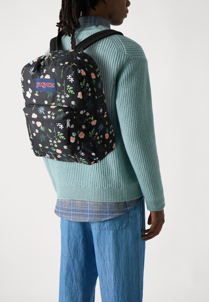 Juodas kuprinės JanSport su gėlių raštu ir keliais skyreliais. Dizainą sudaro rožinės ir baltos gėlės juodame fone.