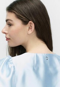 Ljusblå satängblus med puffärmar och en rund metallknappdetalj vid halsringningen, slät textur och mjuk lyster.