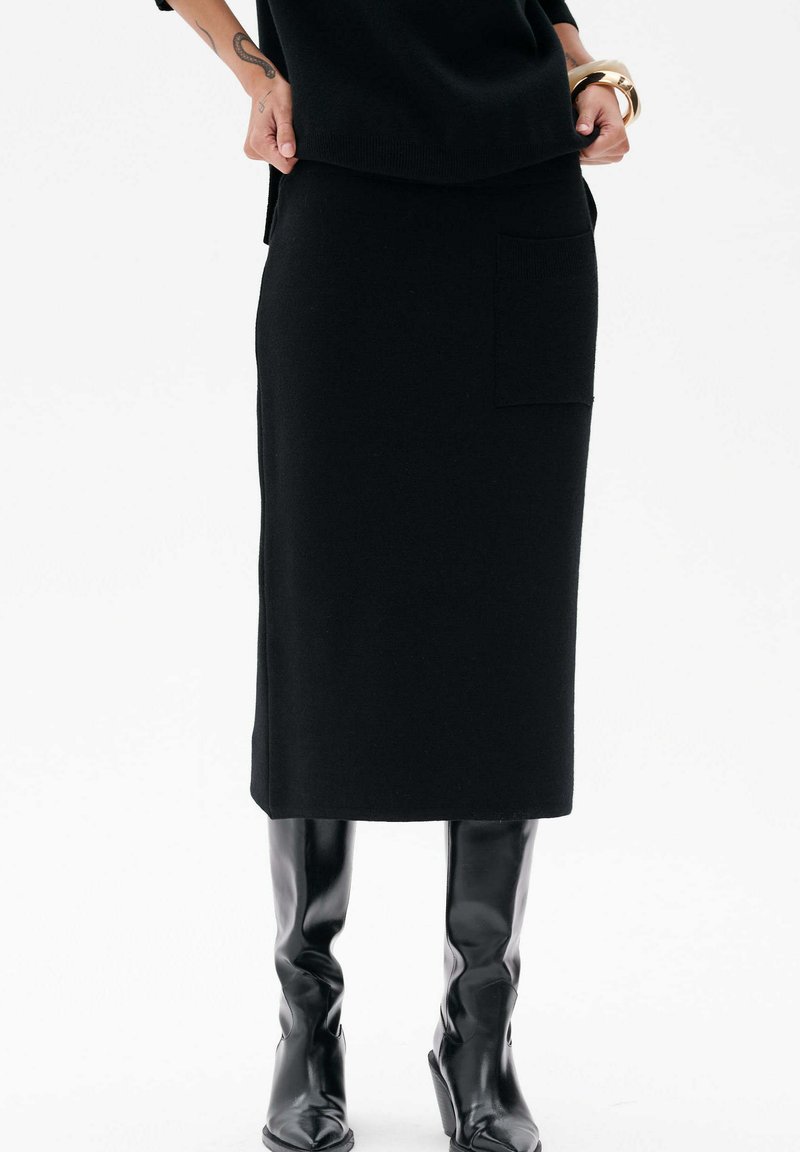 Osoba ubrana w czarną midi spódnicę z boczną kieszenią, czarne botki, czarną bluzkę, złotą bransoletkę i tatuaż na lewym przedramieniu.