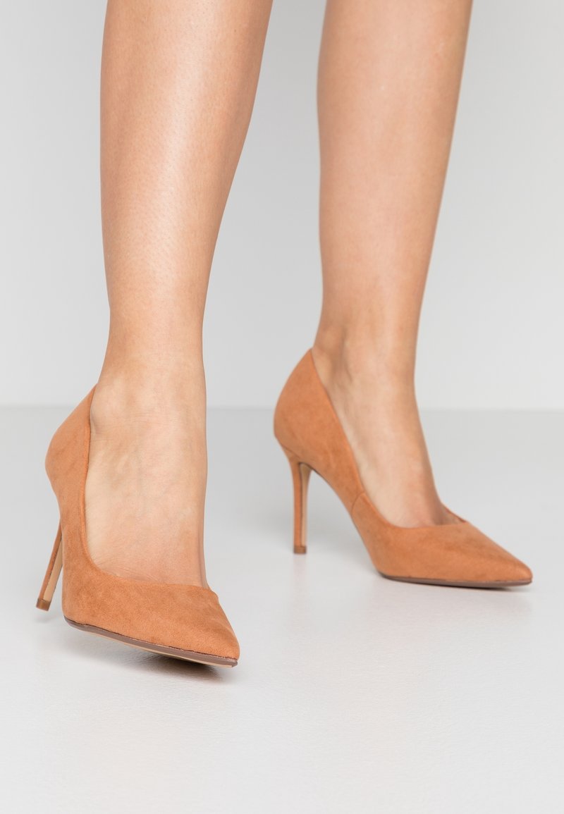Jambes portant des chaussures à talons hauts en daim marron clair à bout pointu, debout sur un sol gris clair sur fond uni.