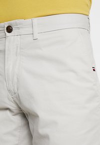 Pantaloni di cotone grigio chiaro con chiusura a bottone, tasche laterali e un piccolo logo tricolore vicino all'orlo.