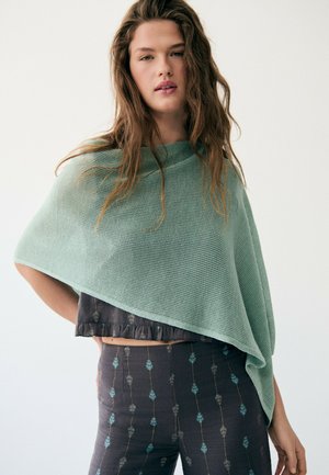 Jeune femme aux longs cheveux portant un poncho tricoté vert clair par-dessus un crop top sombre et un pantalon taille haute à motifs, posant devant un fond blanc.