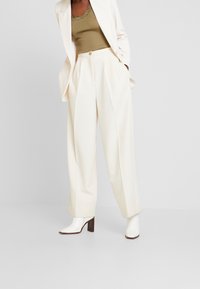 Woman wearing white wide-leg trousers, white heeled ankle boots, a beige ribbed tank top, and a white blazer with hands in pockets.