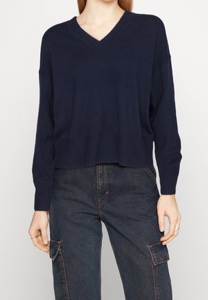 Vrouw die een donkerblauwe V-hals trui met geribbelde zoom en donkergewassen cargobroek met zijzakken draagt, staand tegen een effen achtergrond.