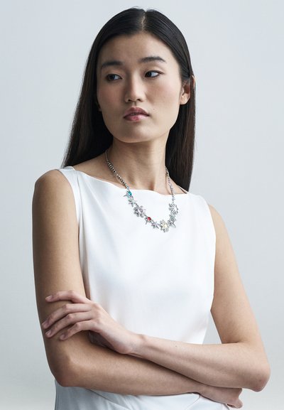 Zilveren bloemenketting met gekleurde accenten met blauwe, rode en gele bloemen. Model draagt een witte mouwloze top en heeft de armen gekruist.