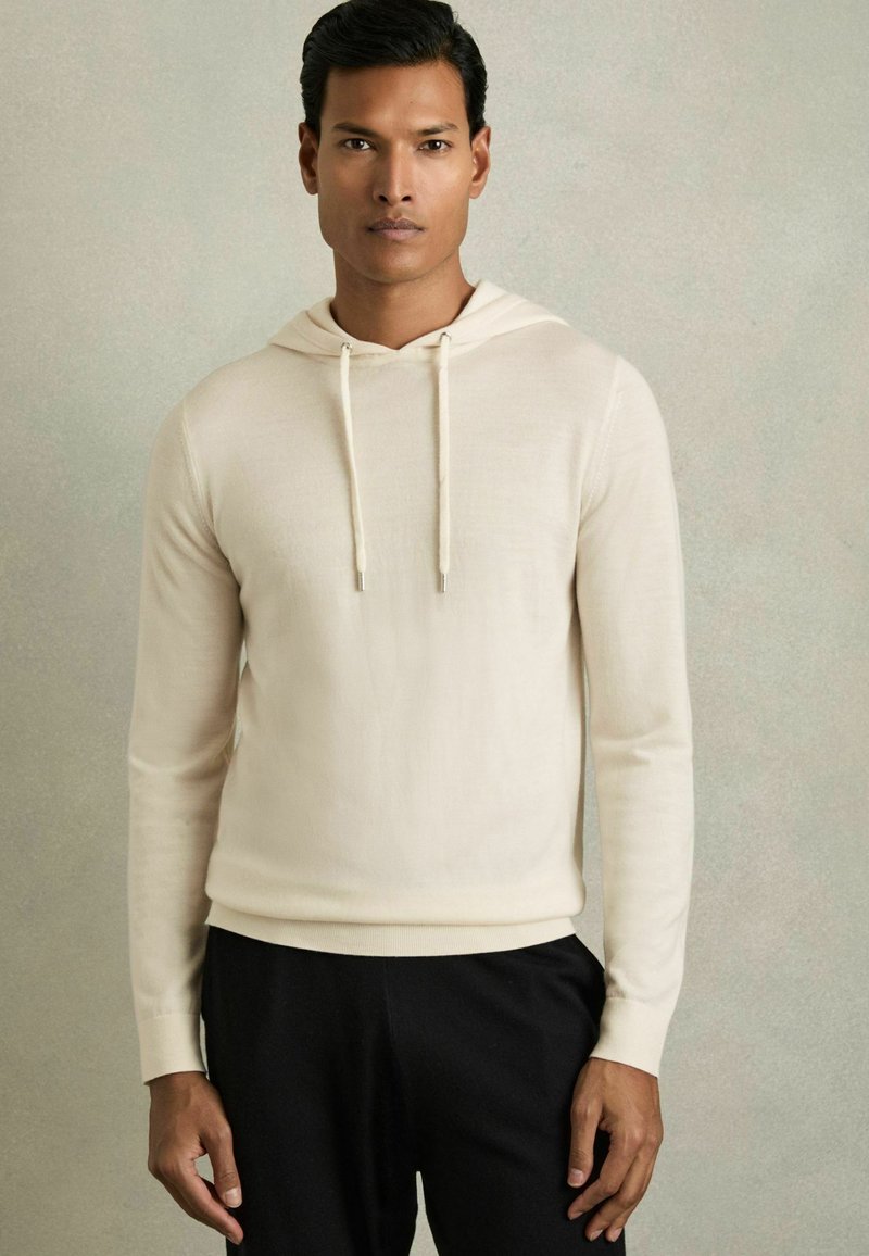 Hellbeige Kapuzenpullover aus weichem Stoff. Er hat eine Kordelzugkapuze, lange Ärmel sowie gerippte Bündchen und Saum. Getragen mit schwarzen Hosen.