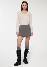 Jasno-beżowy sweter dzianinowy z dekoltem w kształcie litery V, zestawiony z szarym mini spódnicą typu wrap z boczną kieszenią oraz czarnymi botkami na sznurowanie sięgającymi do kolan.