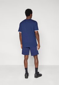 Μπλε σκούρο κοντομάνικο μπλουζάκι με λευκά μανσέτα ως διακοσμητικό στοιχείο, σε συνδυασμό με μπλε σκούρες βερμούδες με πλαϊνές ραφές. Φορεμένο με μαύρα παπούτσια και σκουρόχρωμες κάλτσες.