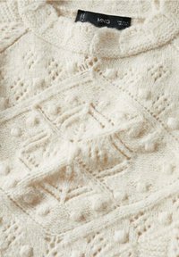 Pull en tricot couleur crème avec des motifs géométriques complexes et des points bouclés, présentant une manche repliée et une étiquette de marque visible.