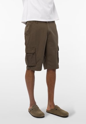 Mann iført brune cargoshorts og taupe slip-on sko, stående med ben synlige fra midje til føtter mot hvit bakgrunn.