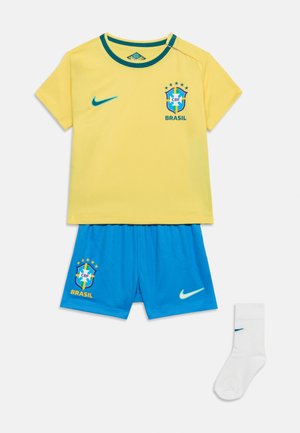 Κίτρινη φανέλα ποδοσφαίρου της Βραζιλίας με μπλε λογότυπο Nike, μπλε σορτς με έμβλημα της Βραζιλίας και λευκά καλτσάκια με το λογότυπο Nike, τοποθετημένα επίπεδα σε λευκό φόντο.