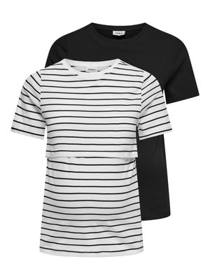 Jeune femme portant une chemise blanche à manches courtes avec des rayures horizontales noires et un pantalon noir à jambes larges, debout les mains dans les poches.