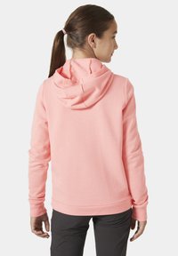 Sudadera con capucha de color rosa claro, fabricada con un material texturizado, con puños y dobladillo acanalados. Presenta un ajuste holgado y se combina con pantalones oscuros.