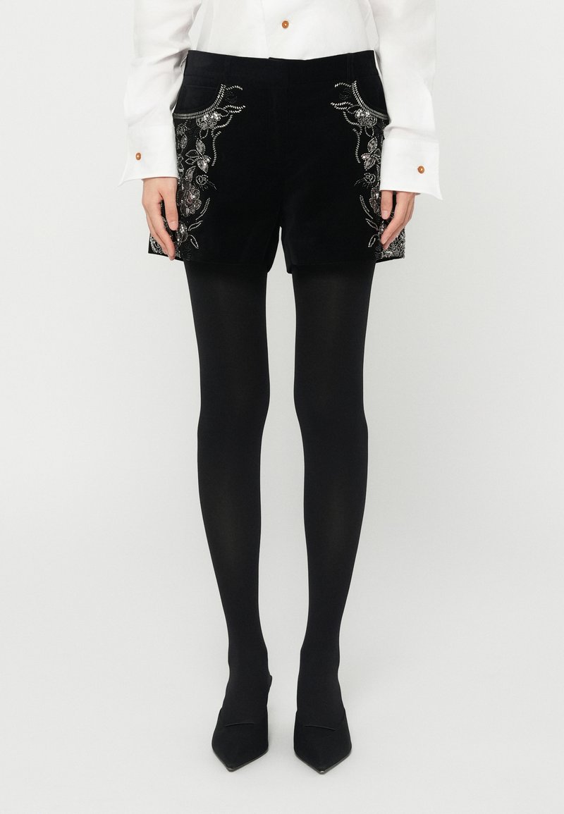 Shorts en velours noir ornés d'une broderie complexe de fleurs et de feuilles sur les poches avant, portés avec des collants noirs et des escarpins �à bouts pointus.