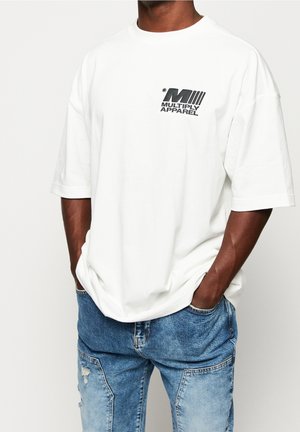 Mężczyzna noszący białą, oversize'ową koszulkę z logo "Multiply Apparel" na piersi oraz niebieskie, podarte dżinsy, z rękami w kieszeniach.