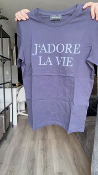 Violeta kokvilnas t-krekls ar īsām piedurknēm un apaļu kaklu, uz kura priekšpuses ir balta uzraksts "J'ADORE LA VIE".