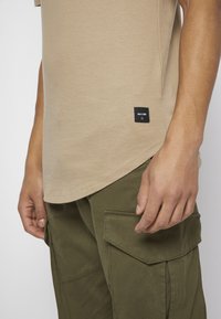 T-shirt bege de manga curta com bainha curved e uma pequena etiqueta preta. Calças cargo verde-oliva com grandes bolsos e costura detalhada.