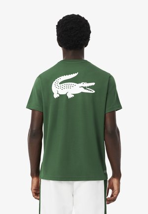 Hombre con camiseta verde con un gran logo blanco de cocodrilo en la espalda, combinada con pantalones cortos blancos que tienen rayas laterales verdes.