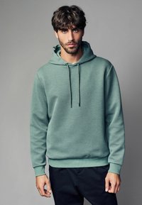 Hoodie u mint zelenoj boji s teksturom, s kapuljačom na šnirc, i rebrastim manžetama, uparen s crnim hlačama. Tkanina ima uzorak dijamanta.