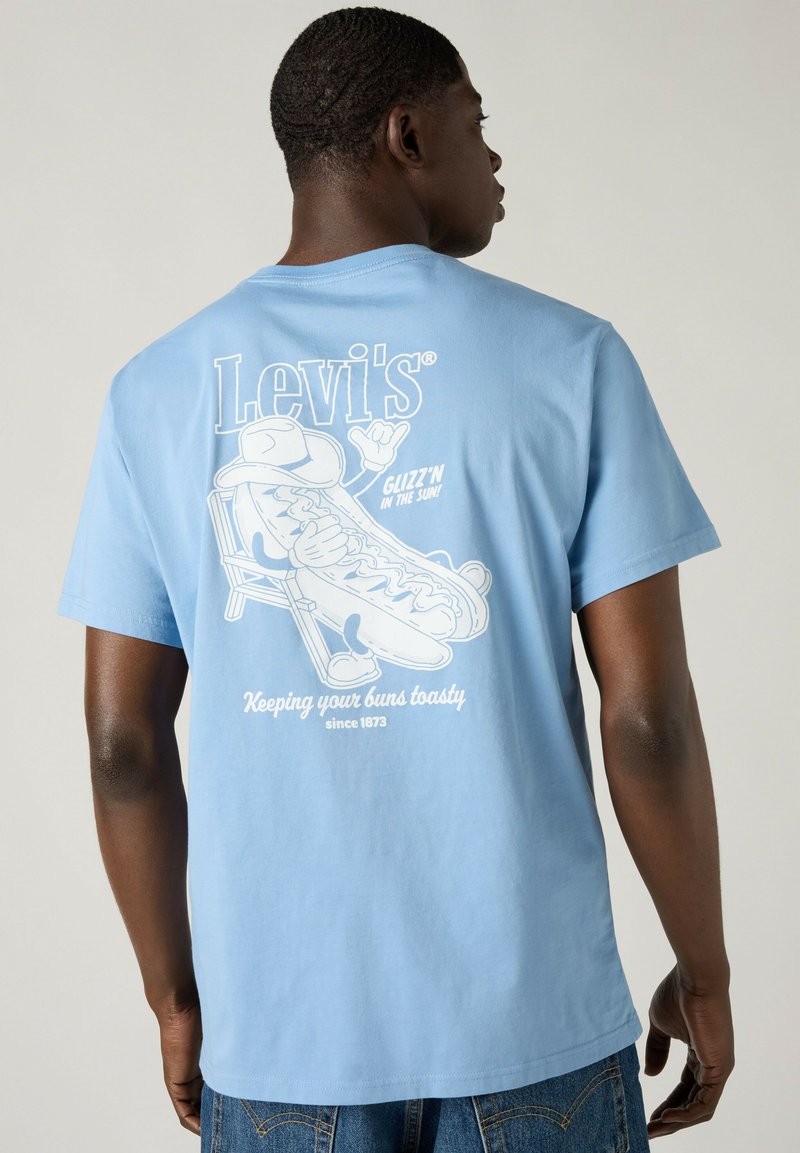 Mann trägt hellblaues Levi's T-Shirt mit einem Cartoon-Hotdog, sitzt auf einem Stuhl und der Text lautet: "Glizz’n in the sun! Keeping your buns toasty."