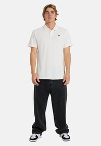 Άσπρο πόλο μπλουζάκι με κοντά μανίκια, γιακά μπροστά και μικρό μαύρο λογότυπο. Συνδυασμένο με άνετο μαύρο τζιν και ασπρόμαυρα αθλητικά παπούτσια.