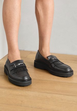 Mocassins à plateforme en cuir noir avec franges et détail de boucle en métal, portés pieds nus sur un sol en bois.