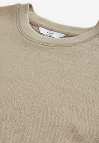 Sweatshirt bej cu un finisaj texturat, având un guler rotund și un material moale. Eticheta spune "NEXT RELAXED FIT."