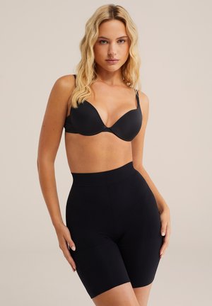 Blondínka v čiernej podprsenke a čiernych sťahovacích šortkách s vysokým pásom stojaca s rukami položenými pri bokoch na béžovom pozadí.