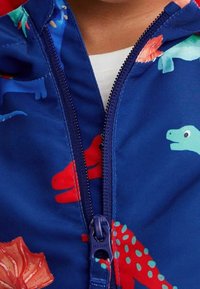 Veste zippée bleue avec des motifs de dinosaures en rouge et vert. Elle présente une texture lisse et un zip à l'avant pour la fermeture.