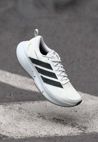 Λευκό αθλητικό παπούτσι με τρεις μαύρες ρίγες, ελαφρύ σχεδιασμό, υφασμάτινο επάνω μέρος, επενδεδυμένο κολάρο και μαξιλαρωμένη σόλα, εμφανιζόμενο στον αέρα.