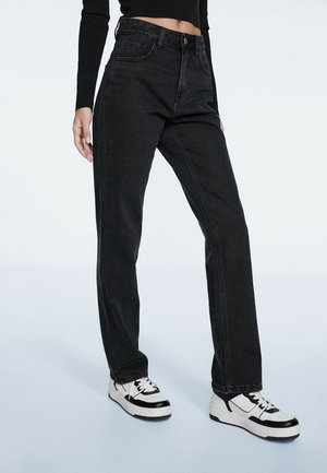 Black high-waisted jeans with a straight fit, denim material, featuring front pockets and a button closure. Paired with white sneakers.