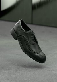 Chaussure habillée en cuir noir avec des perforations, un laçage et un bout arrondi. Semelle texturée de forme minimaliste.