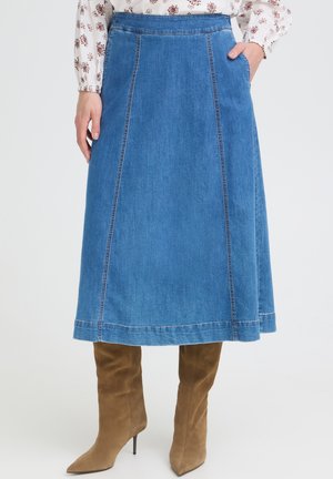 Frau trägt einen knielangen blauen Jeansrock mit vorderen Nähten, braune Wildlederstiefel mit hohen Absätzen und eine weiße Blumenbluse.