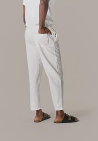 Persona de pie, vestida con pantalones de lino blancos con cintura elástica y bolsillo parche, combinados con sandalias negras de doble tira sobre un fondo neutro.